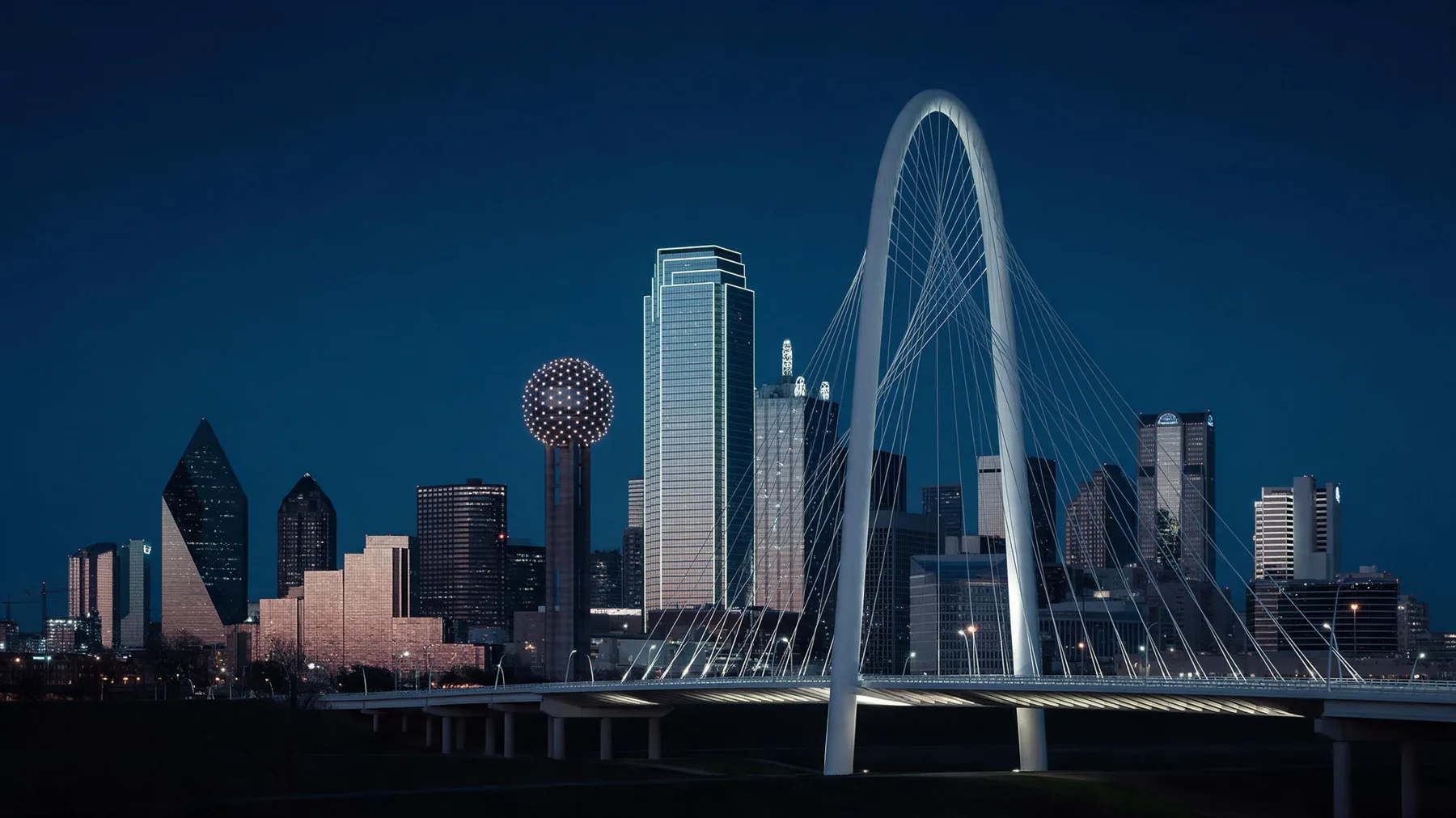 Dallas, Texas downtown skyline at blue hour behind a lit arch bridge